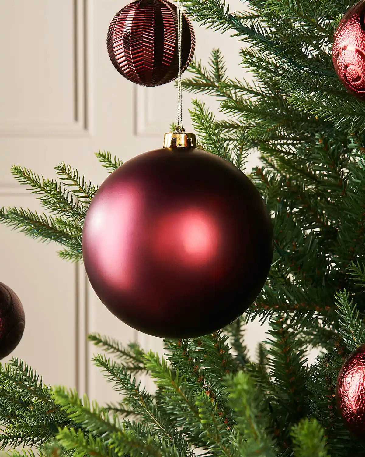 Matt burgundy 15cm oversized bauble on a Christmas tree branch with 4 smaller burgundy baubles in the background 