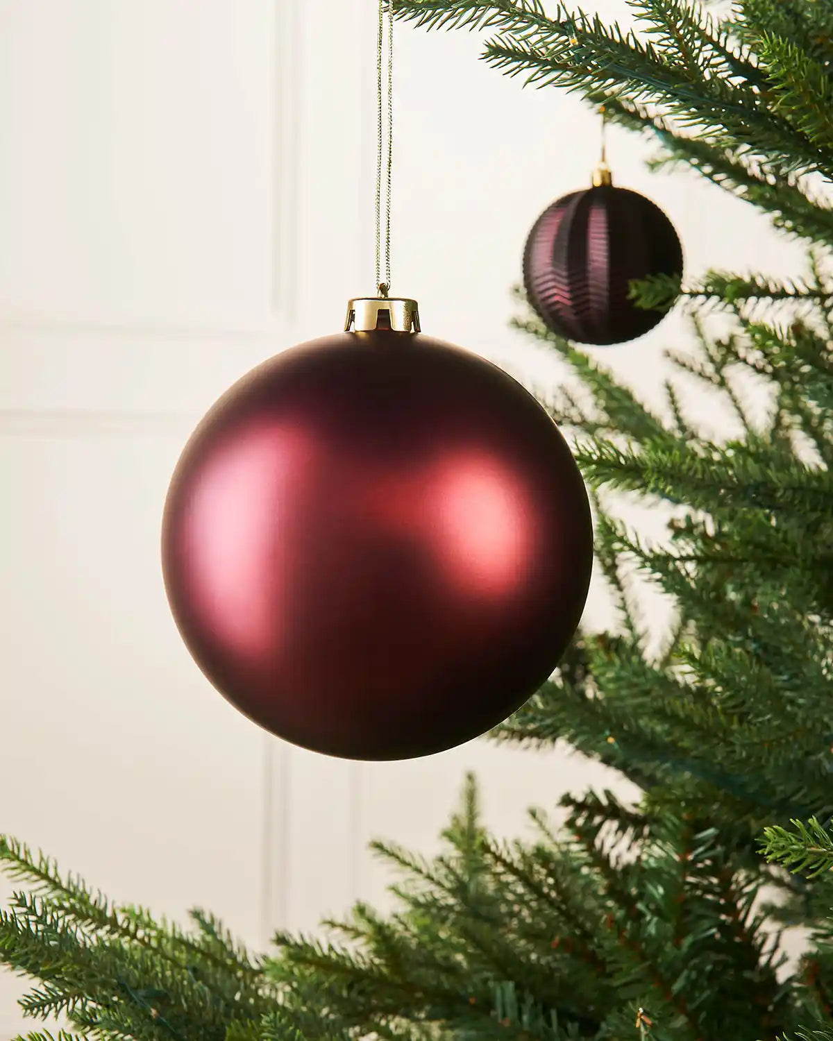 Matt burgundy 15cm oversized bauble on a Christmas tree branch with a smaller burgundy bauble in the background