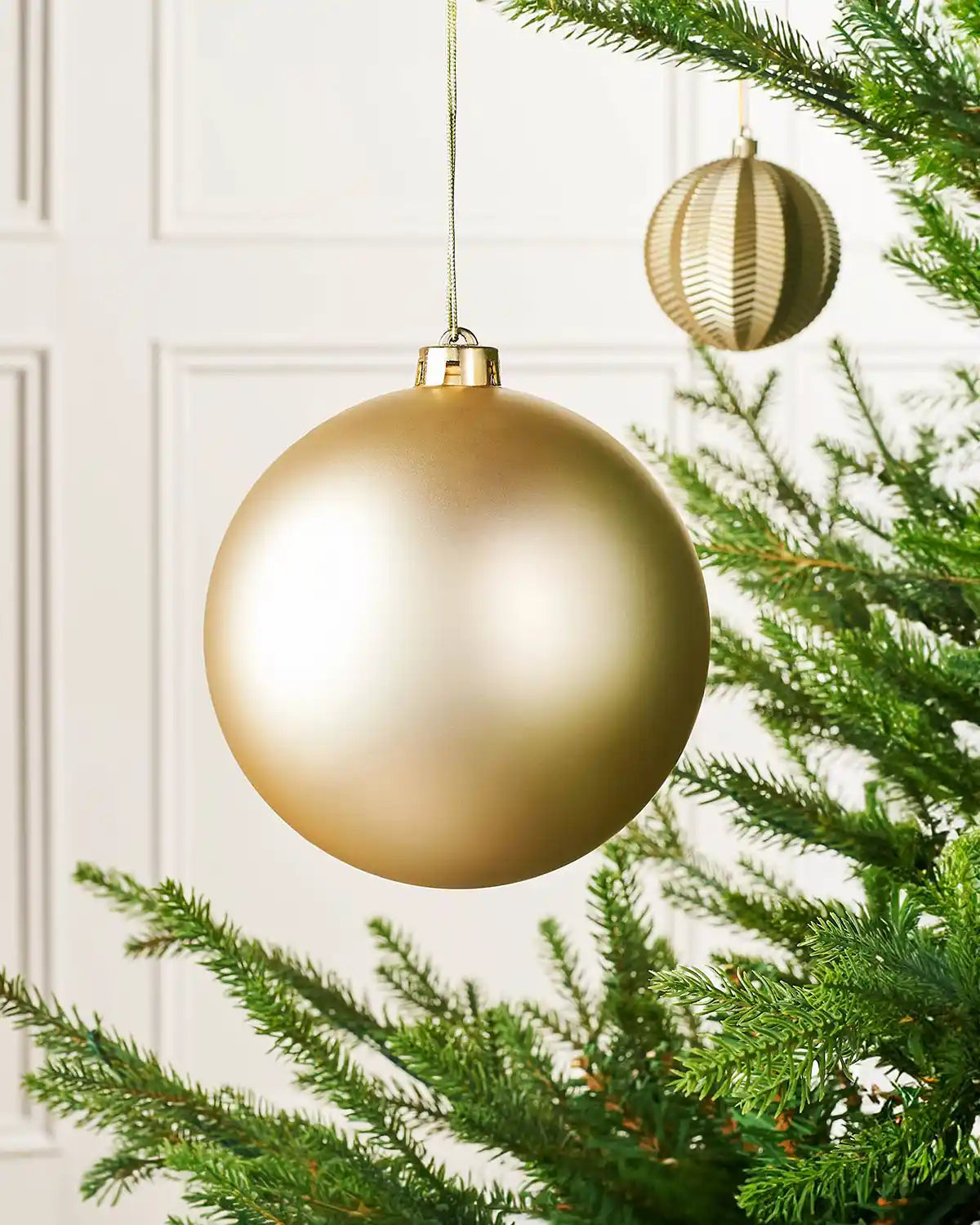 Matt gold 15cm oversized bauble on a Christmas tree branch with a smaller gold bauble in background