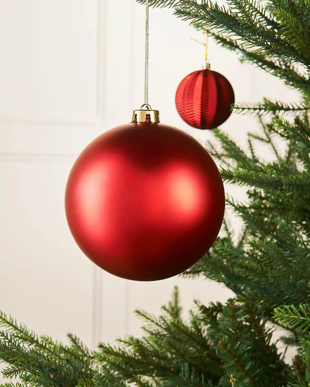Matt red 15cm oversized bauble on a Christmas tree branch with a smaller red bauble in the background