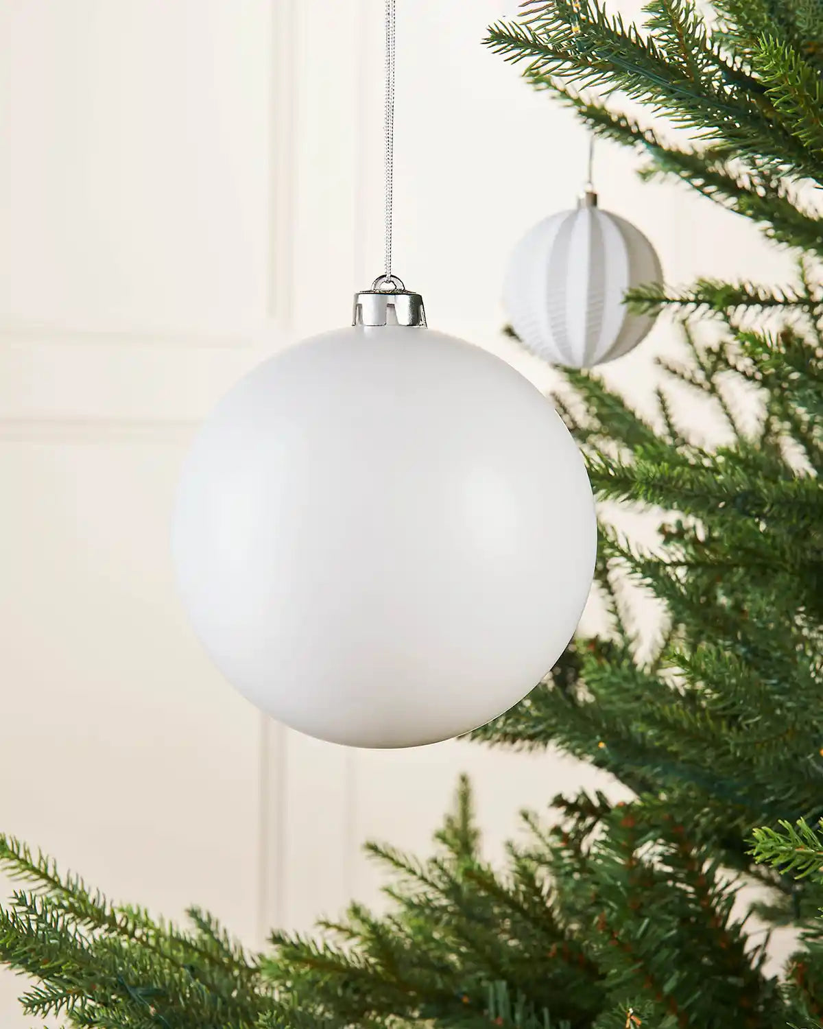 Matt white 15cm oversized bauble on a Christmas tree branch with a smaller white bauble in background