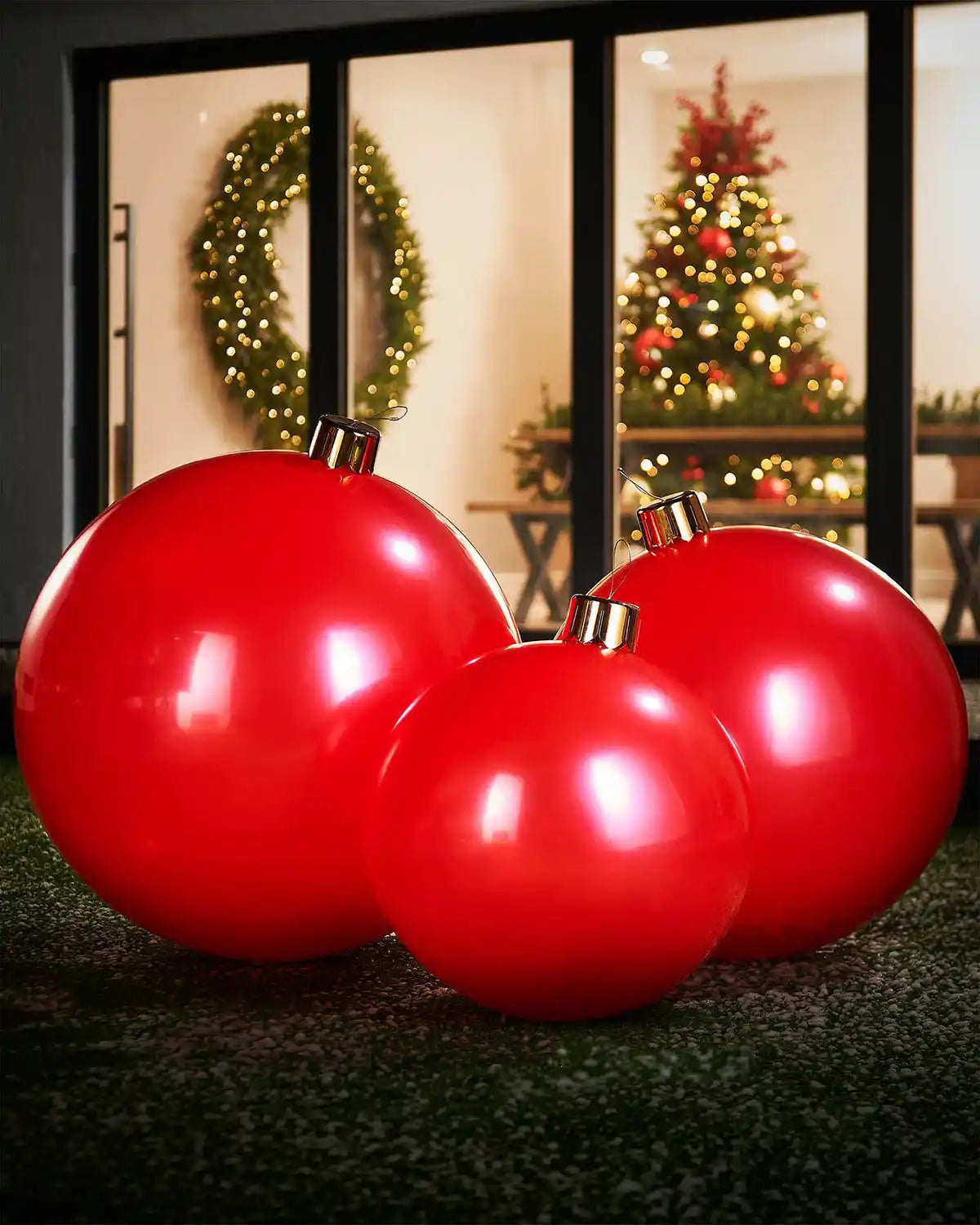 Medium, large and extra large Christmas  red inflatable baubles on a grassy surface with a festive indoor background.
