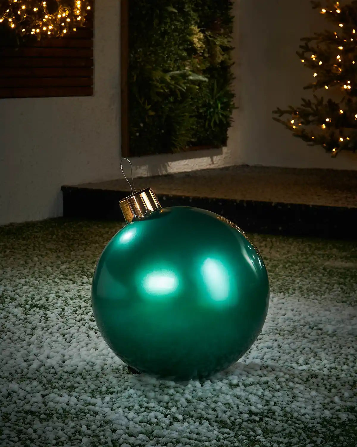 Medium sized green inflatable bauble on a snowy garden floor with a decorated Christmas tree and wreath in background 
