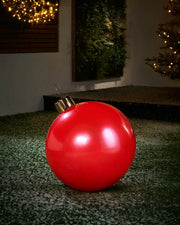 Medium sized red inflatable bauble on a snowy garden floor with a decorated Christmas tree and wreath in background 