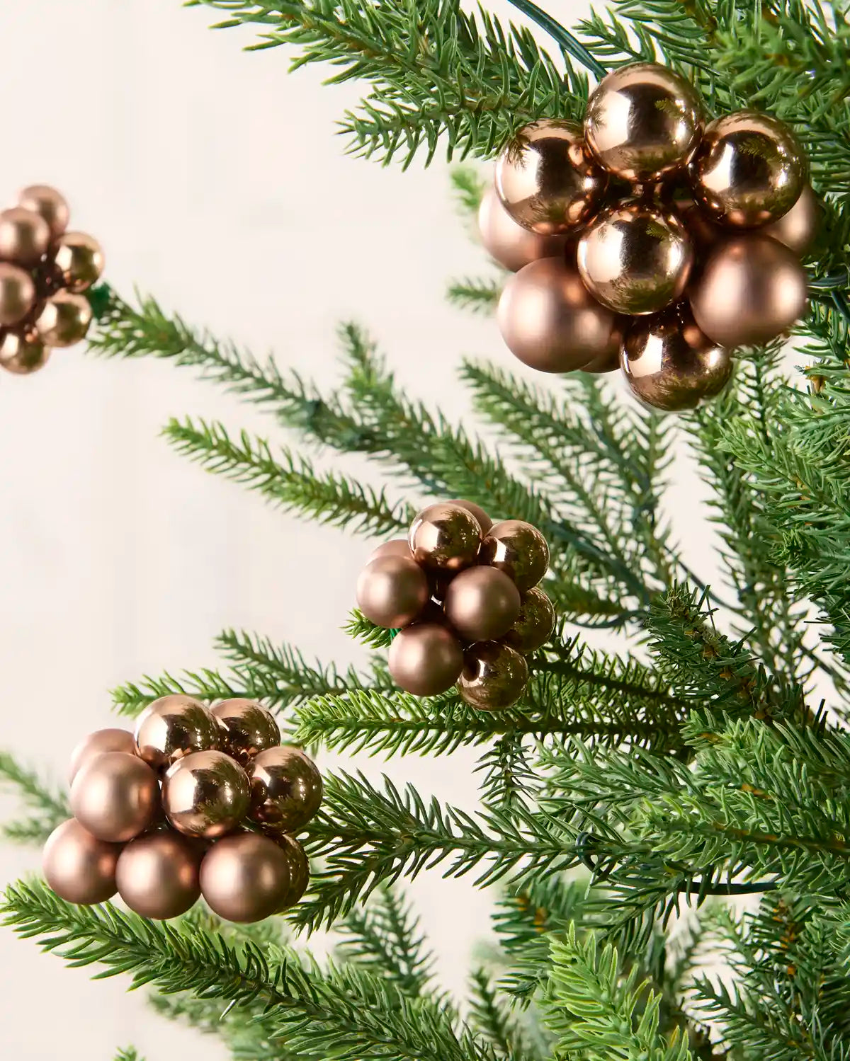 Metallic Mocha berry cluster Christmas tree ornaments hanging on green pine branches against a soft off-white background.
