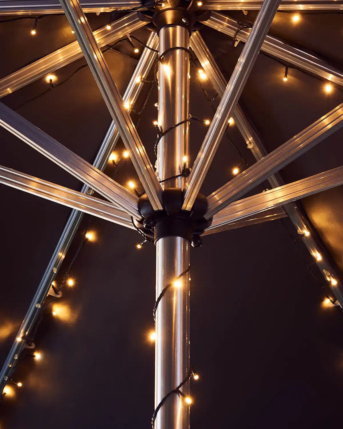 Metallic outdoor umbrella pole with warm white string lights against a dark background