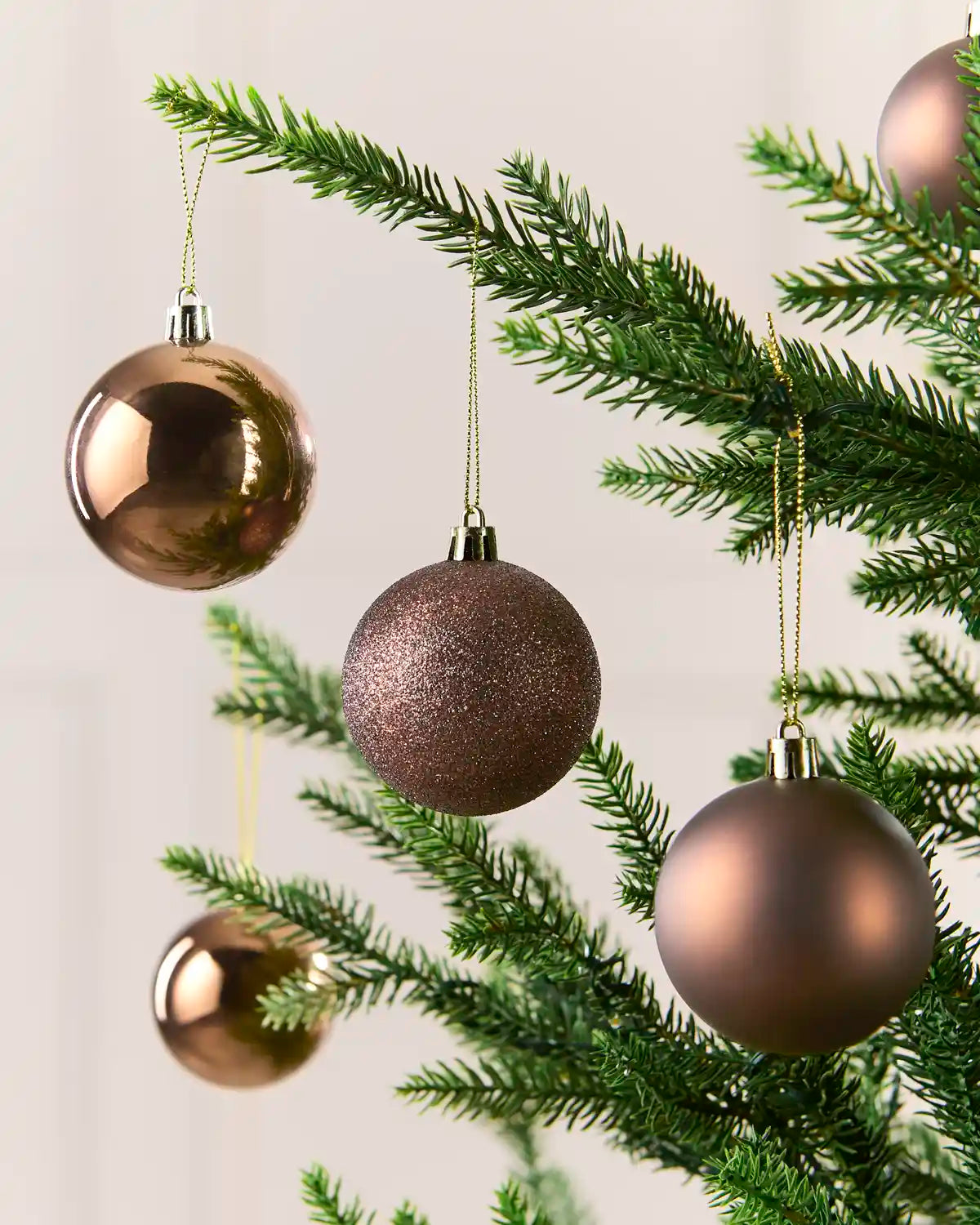 Mocha Christmas tree ornaments in three finishes—shiny, glittery, and matte—hanging on green pine tree branches against a soft off-white background.
