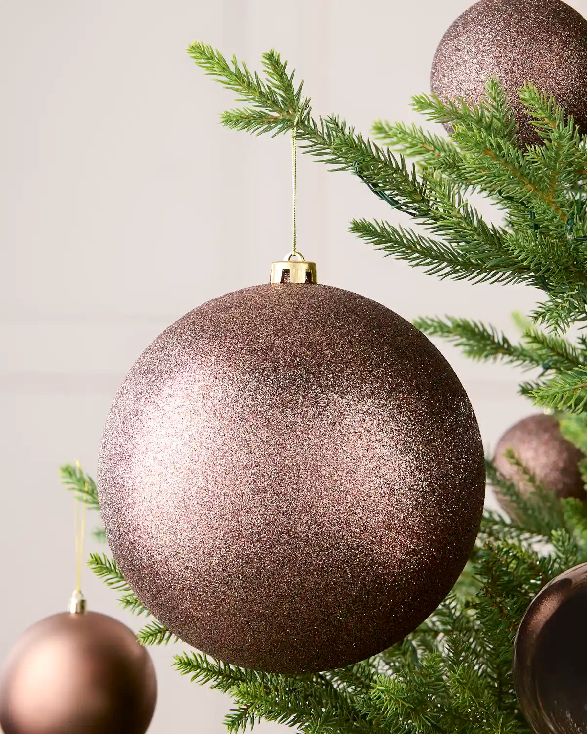 Mocha oversized glitter bauble hanging on a Christmas tree with 4 smaller baubles in the background against a cream wall