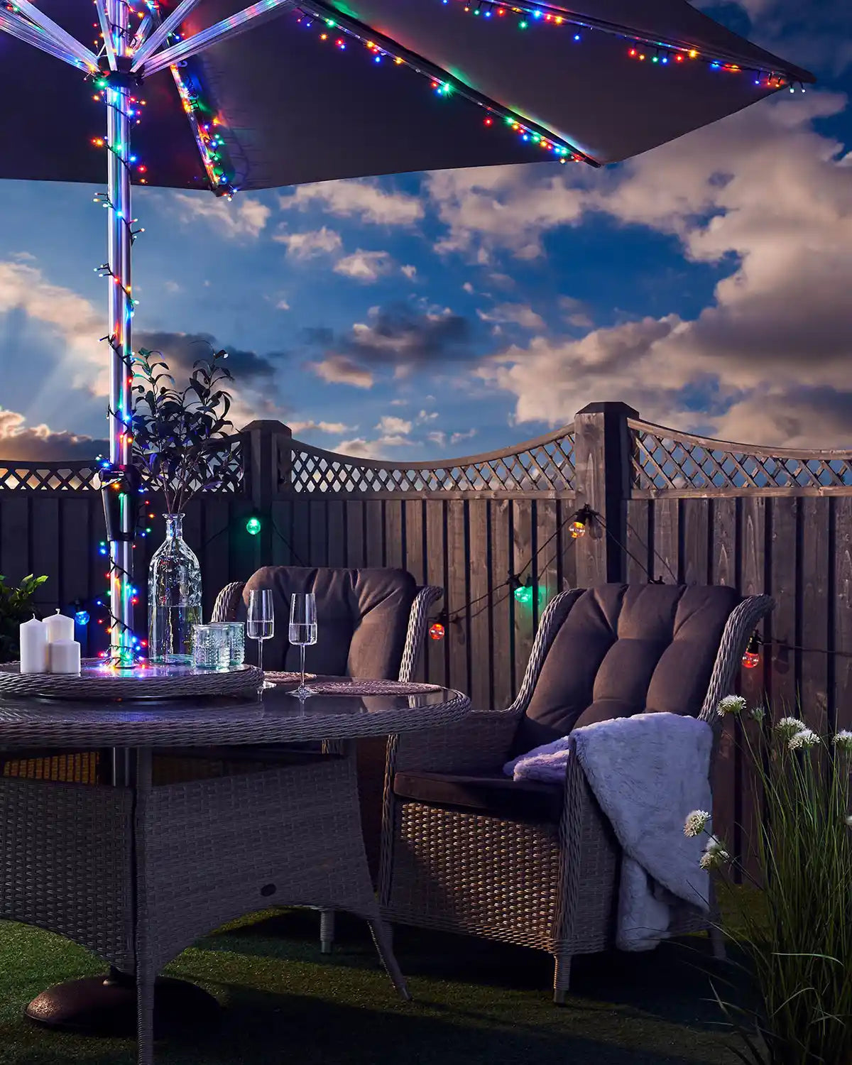 Multi colour LED fairy lights on an umbrella in a garden with table, chairs and glasses of water against a cloudy blue sky 