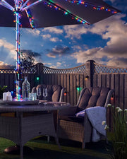 Multi colour LED fairy lights on an umbrella in a garden with table, chairs and glasses of water against a cloudy blue sky 