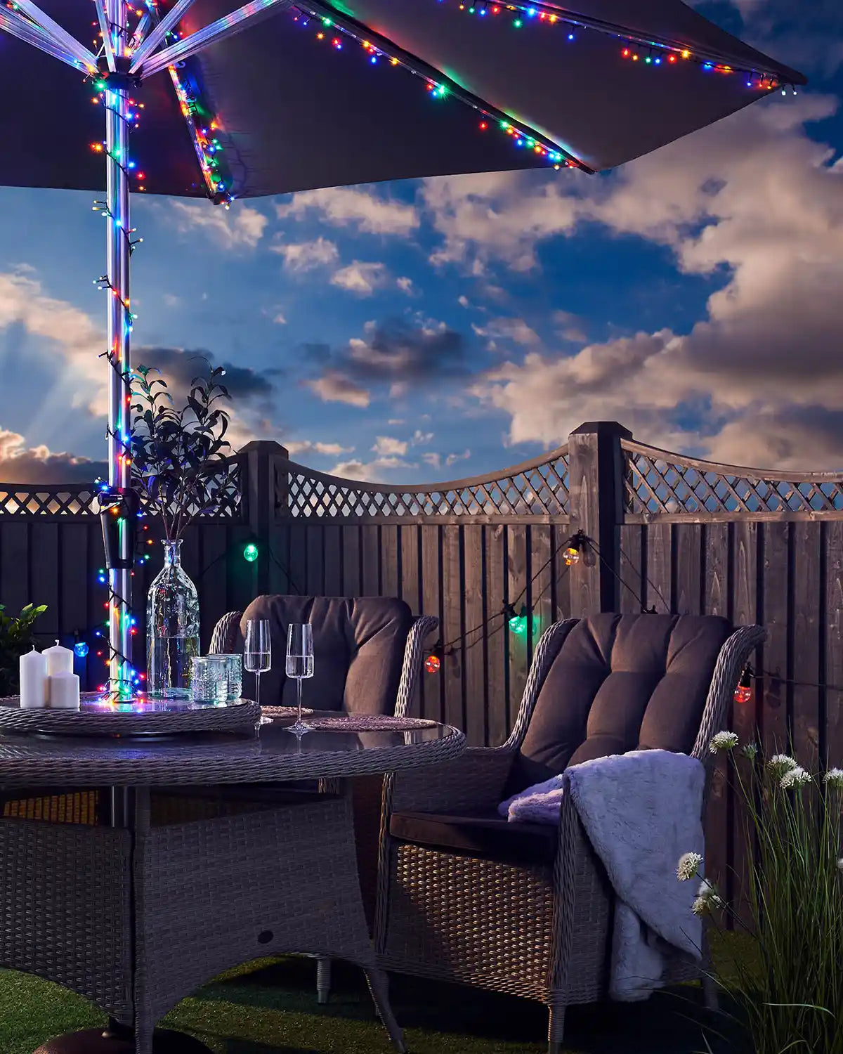 Multi coloured LED fairy lights on an umbrella with garden furniture and cloudy blue sky