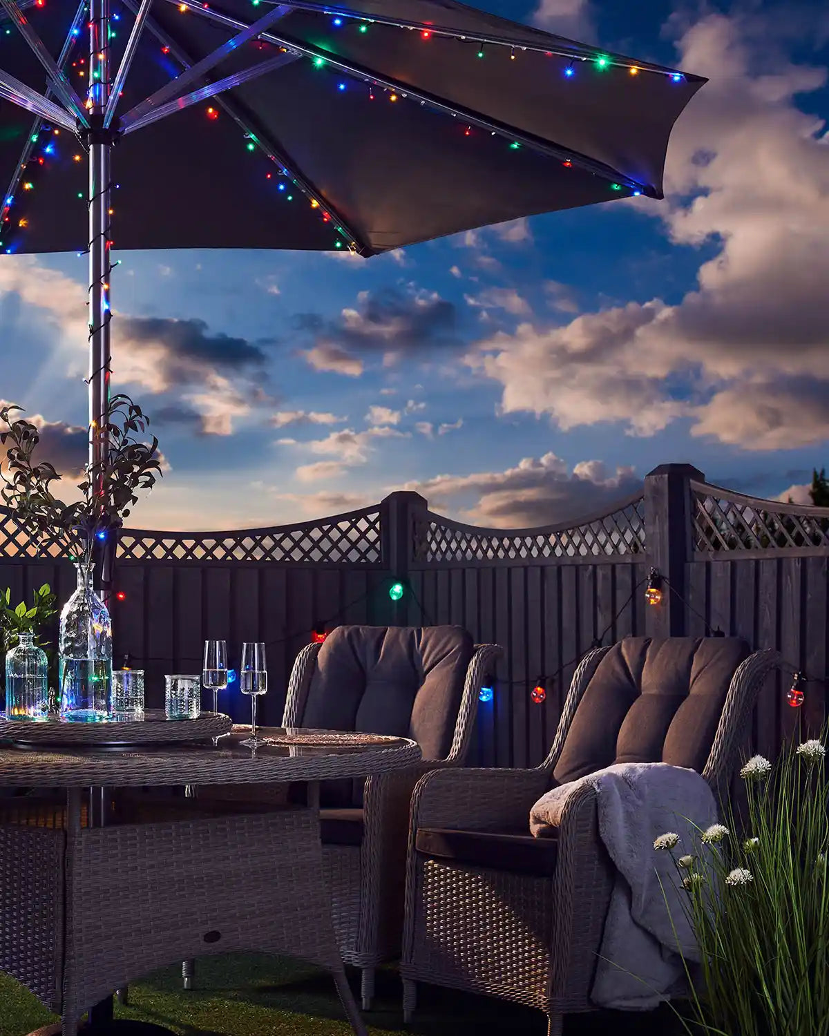 Multi coloured fairy lights wrapped around an umbrella post in a garden with a cloudy blue sky