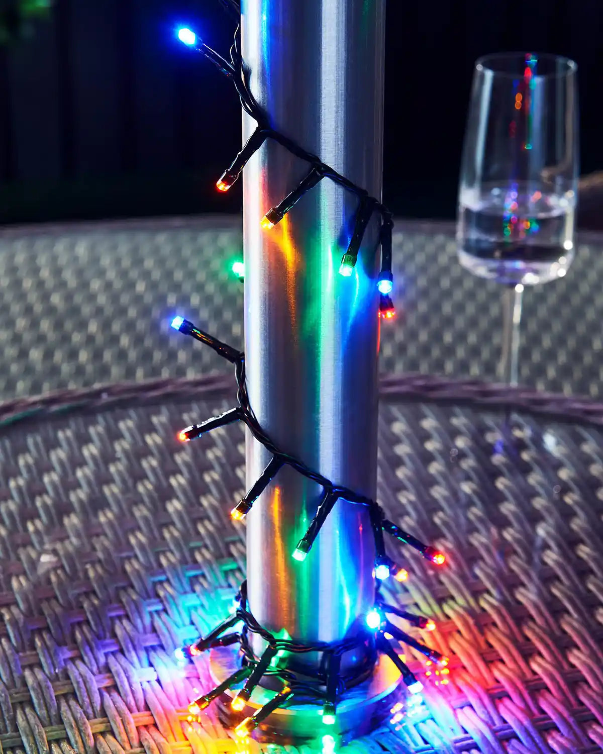 Multicoloured string lights wrapped around a metallic object on a textured surface with a glass in the background.