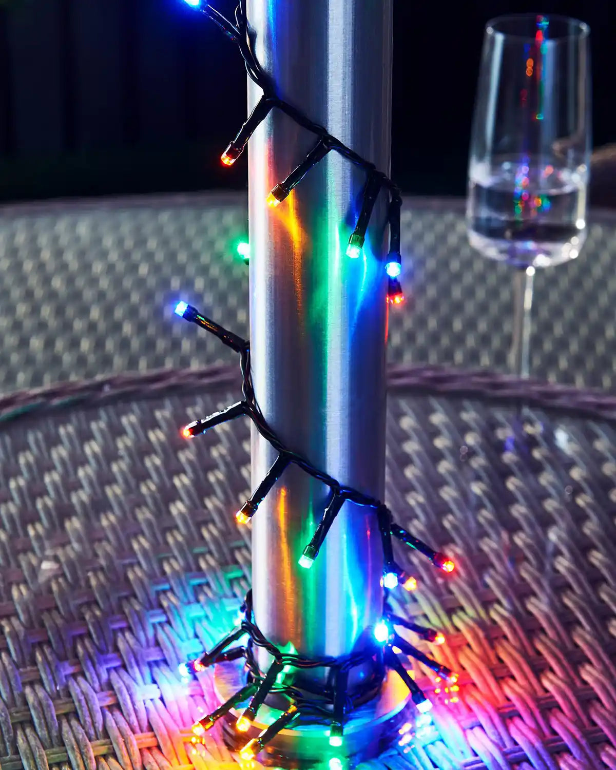 Multicoloured string lights wrapped around a metallic pole with a glass of water on a textured surface.