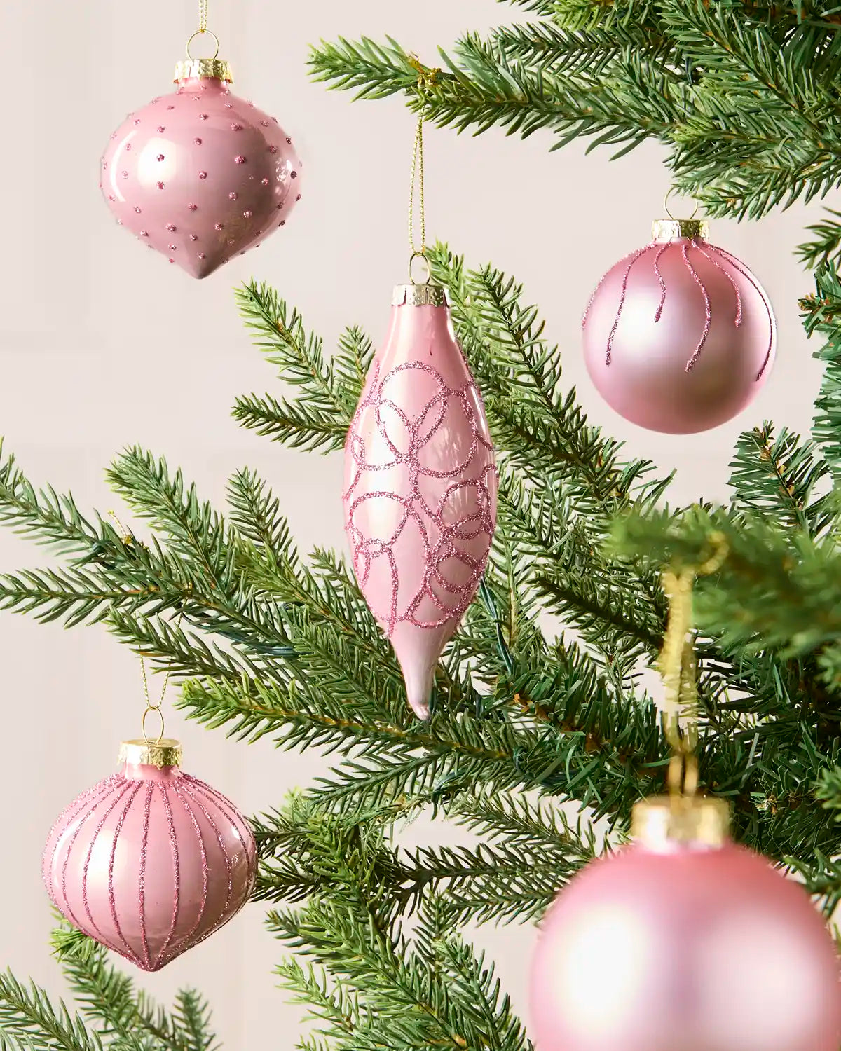 Multiple pink glass baubles hanging from Christmas tree branches in teardrop, onion and classic round shapes, with a blurred cream background.