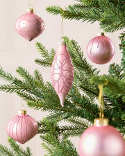Multiple pink glass baubles hanging from Christmas tree branches in teardrop, onion and classic round shapes, with a blurred cream background.