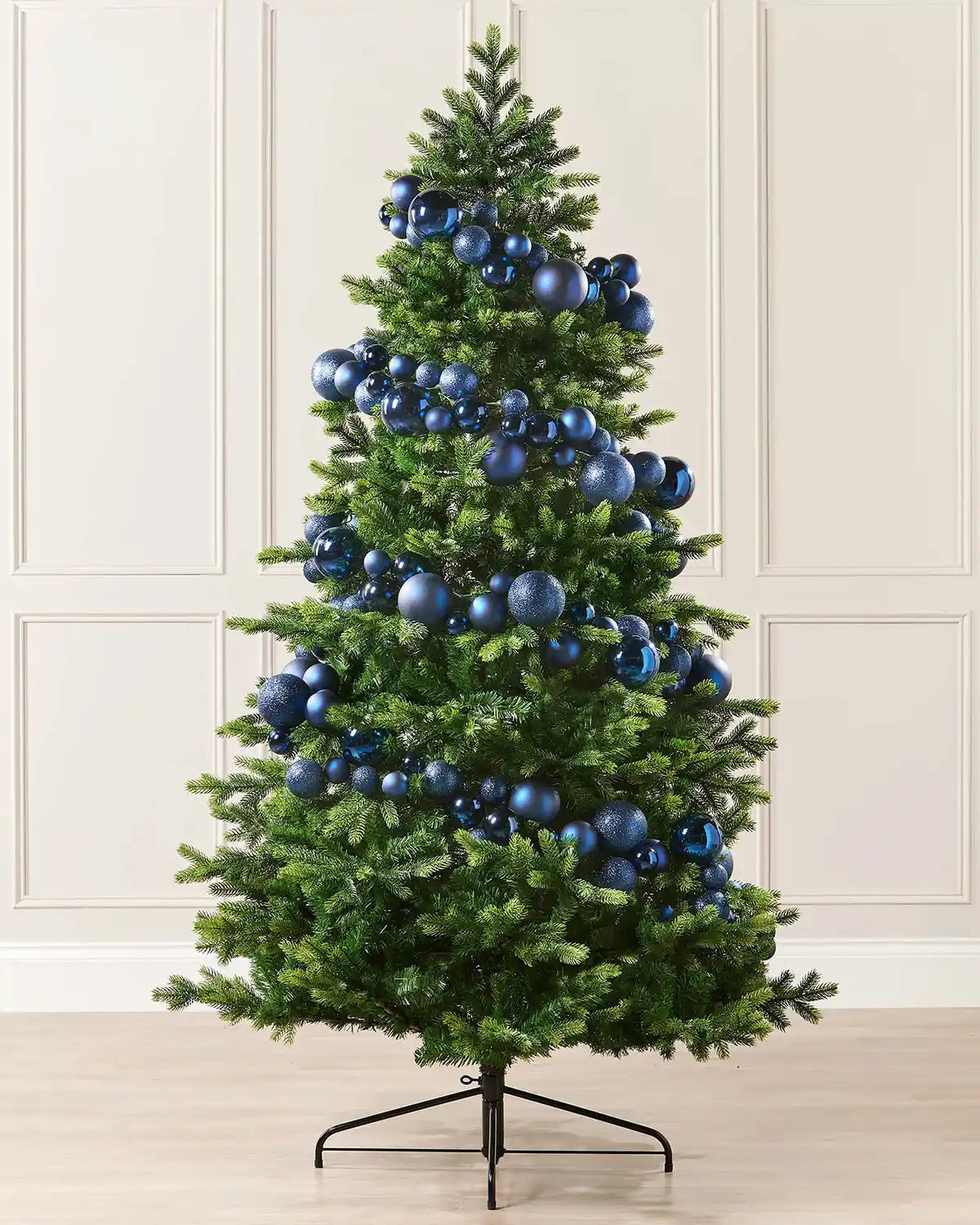 Navy blue bauble garland wrapped around a Christmas tree against a cream panel wall and a wooden floor 
