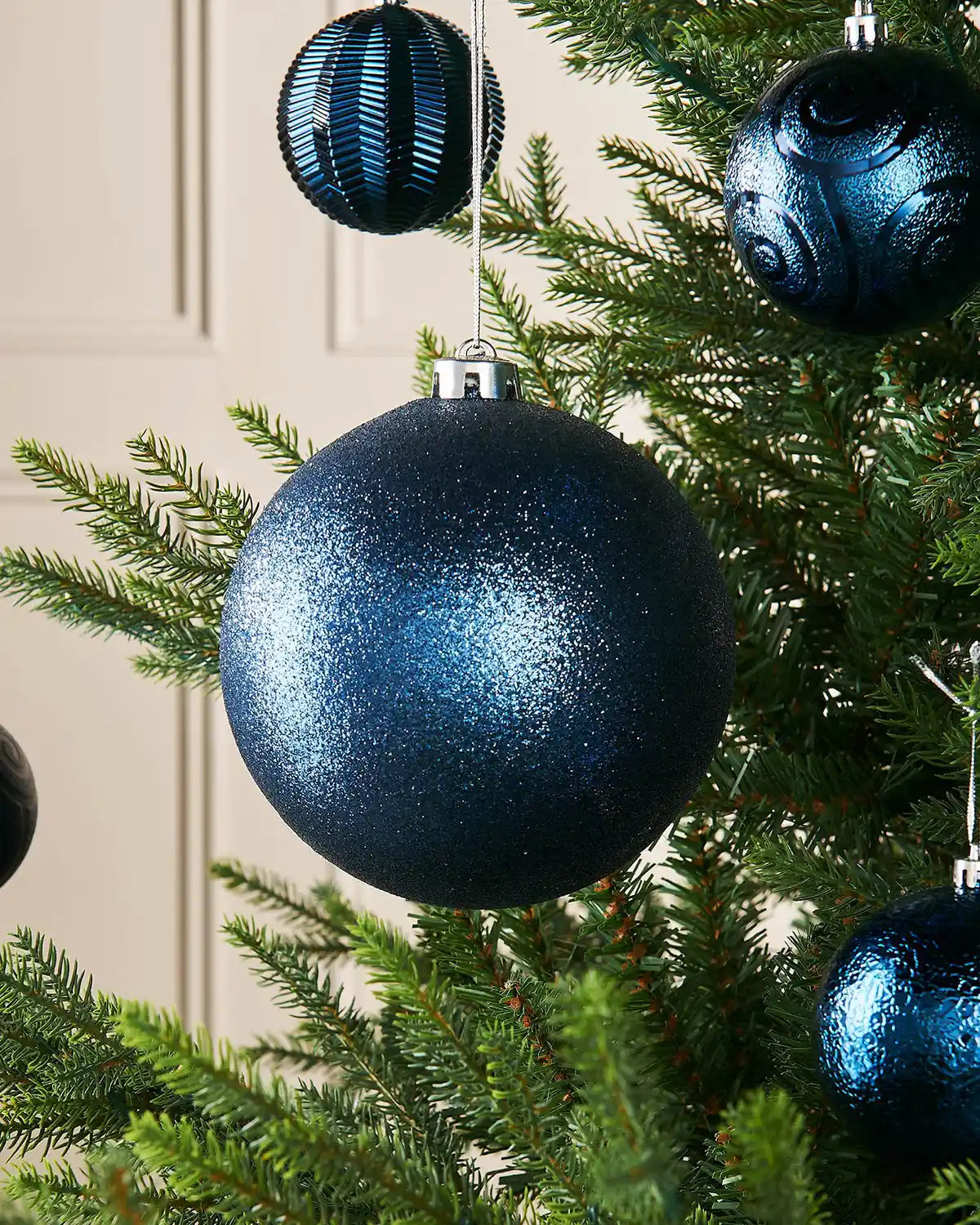 Navy blue glitter oversized 15cm bauble on a Christmas tree branch with 4 smaller baubles in the background