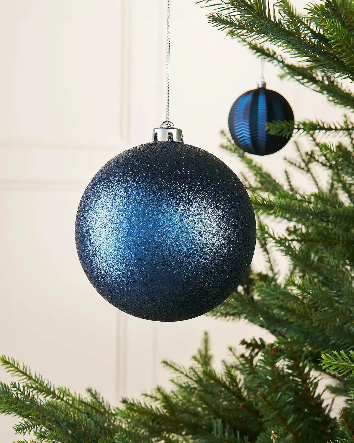Navy blue glitter oversized 15cm bauble on a Christmas tree branch with a smaller bauble in the background