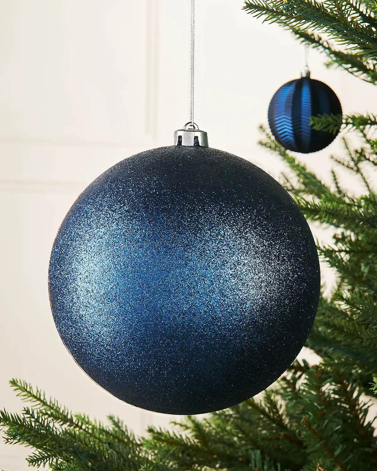 Navy blue glitter oversized bauble on a Christmas tree branch with a smaller navy blue bauble in background