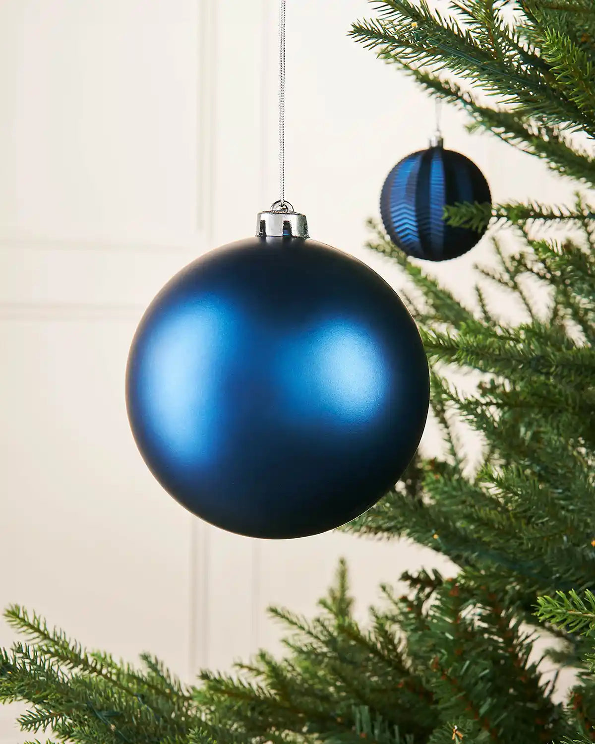 Navy blue matt oversized 15cm bauble on a Christmas tree branch with a smaller blue bauble in background