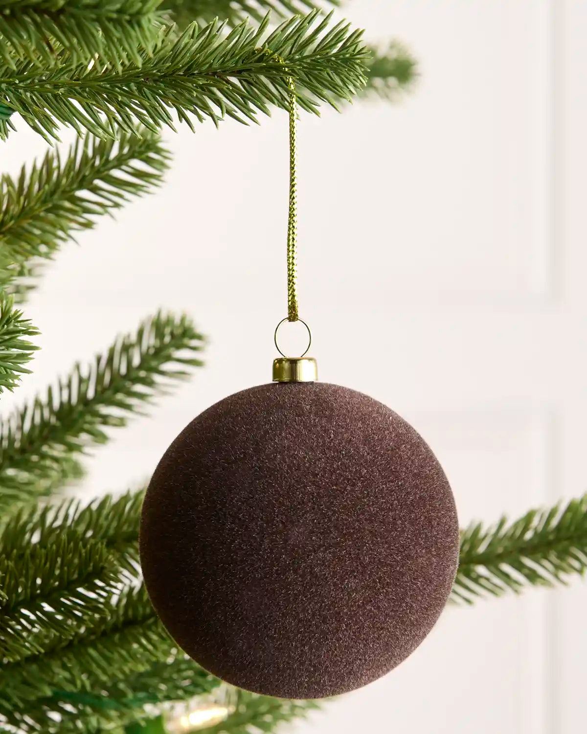 One Espresso coloured velvet Christmas tree bauble hanging from gold chains on green pine tree branches against a light background.