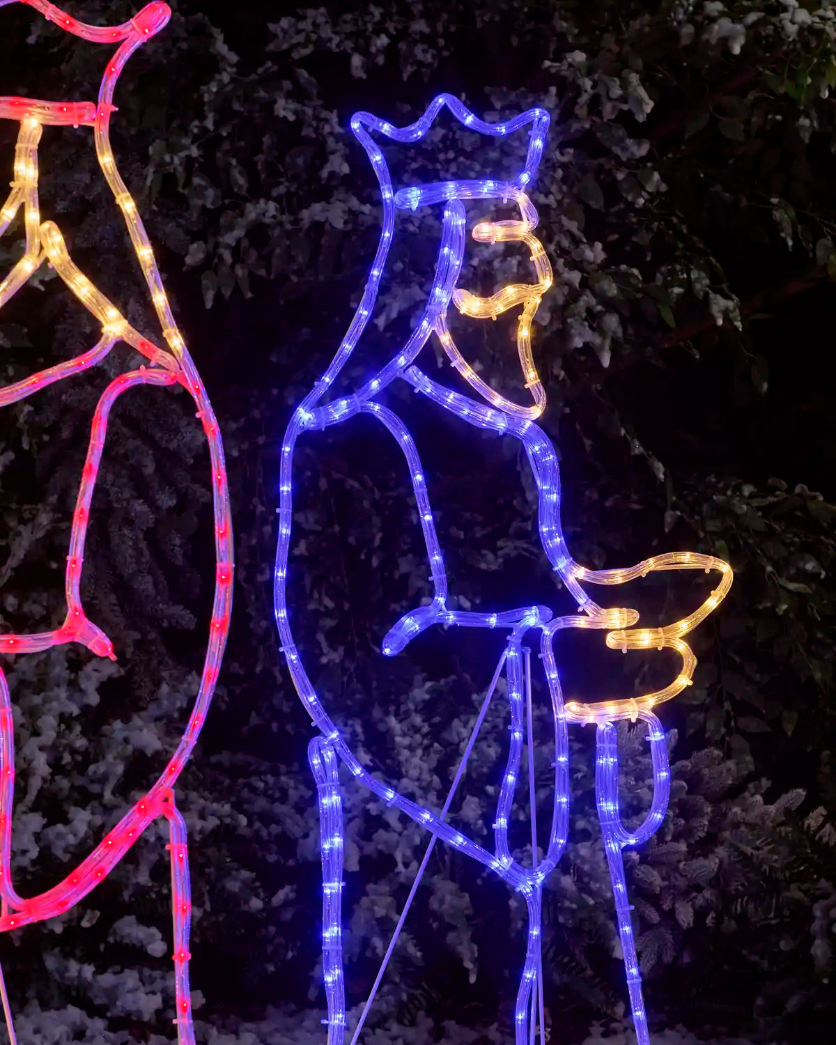 Outdoor Christmas wise man decoration in the shape of a crowned figure with a long beard, outlined in blue LED lights with warm yellow accents, set against a dark night background with snow on the ground.