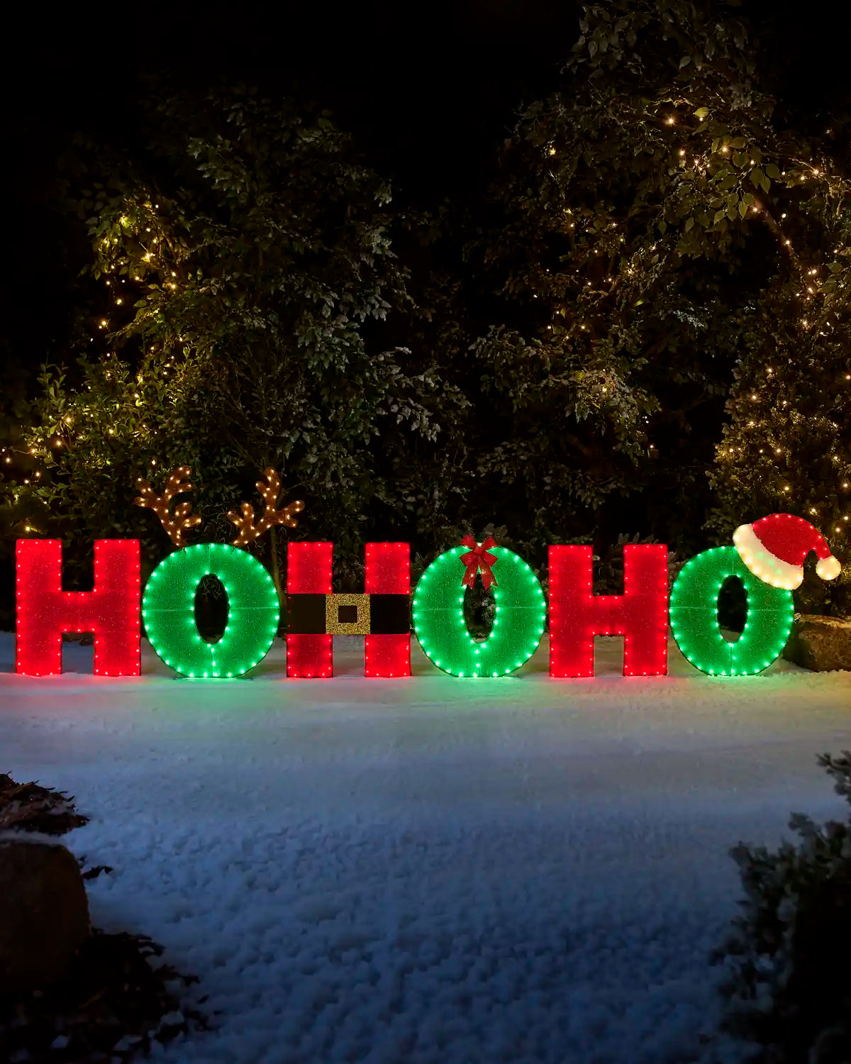 Outdoor HOHO holiday light decoration spelling out 'HO HO HO' with green wreath-style O's, red block letters, a small Santa hat on the right, and a gold accent between the middle O's, displayed in a snowy setting at night with trees lit by warm string lights.