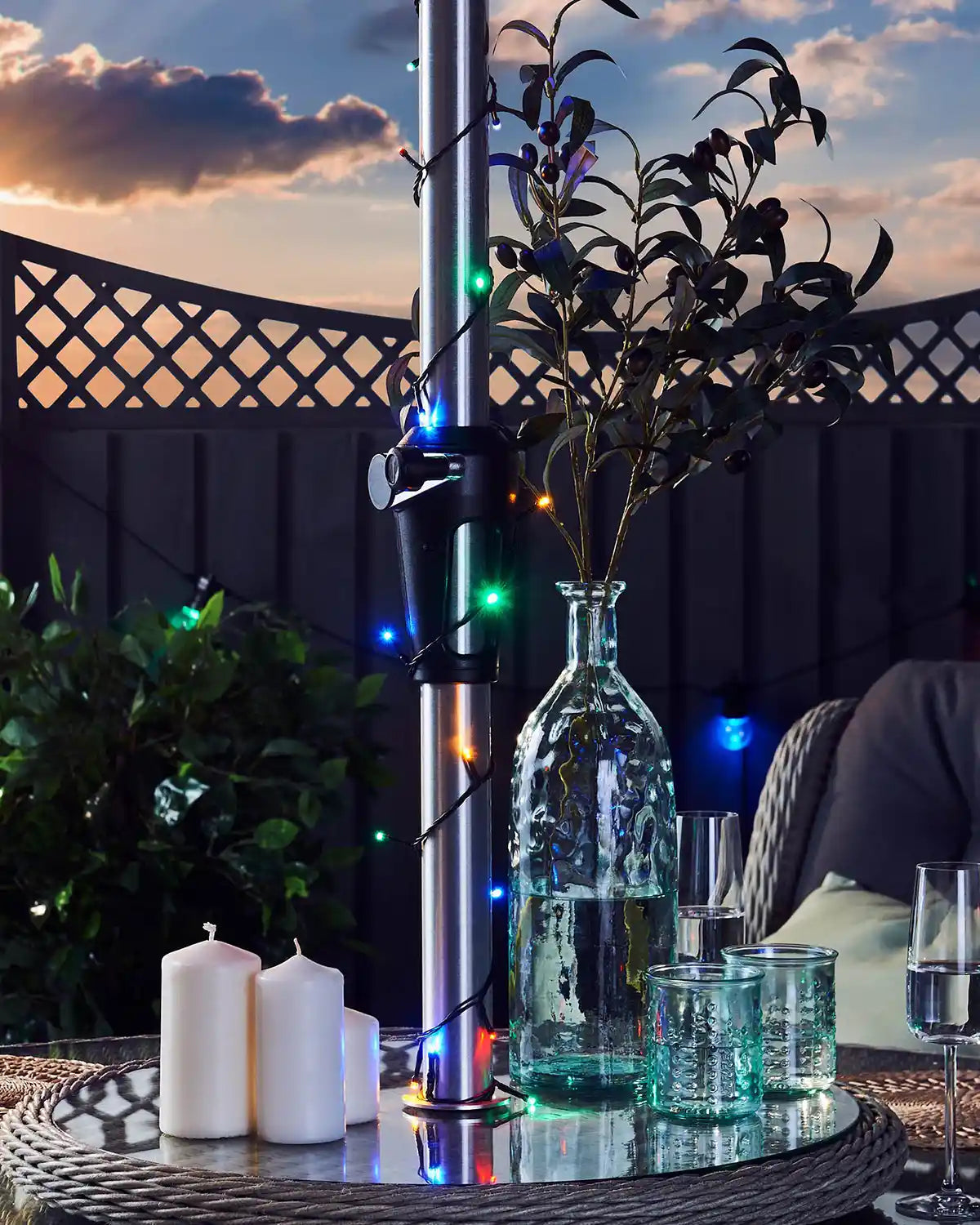 Outdoor evening setting with multi colour string lights wrapped around umbrella pole, candles, glasses, and decorative plants on a table.