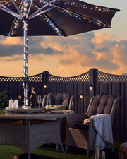 Outdoor garden furniture setting with a warm white and ice white fair light lit umbrella, table, chairs, and sunset sky.