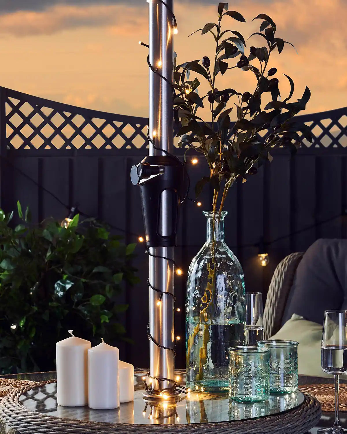 Outdoor garden setting with a table featuring a vase, warm white string lights, candles, and glasses against a sunset backdrop.