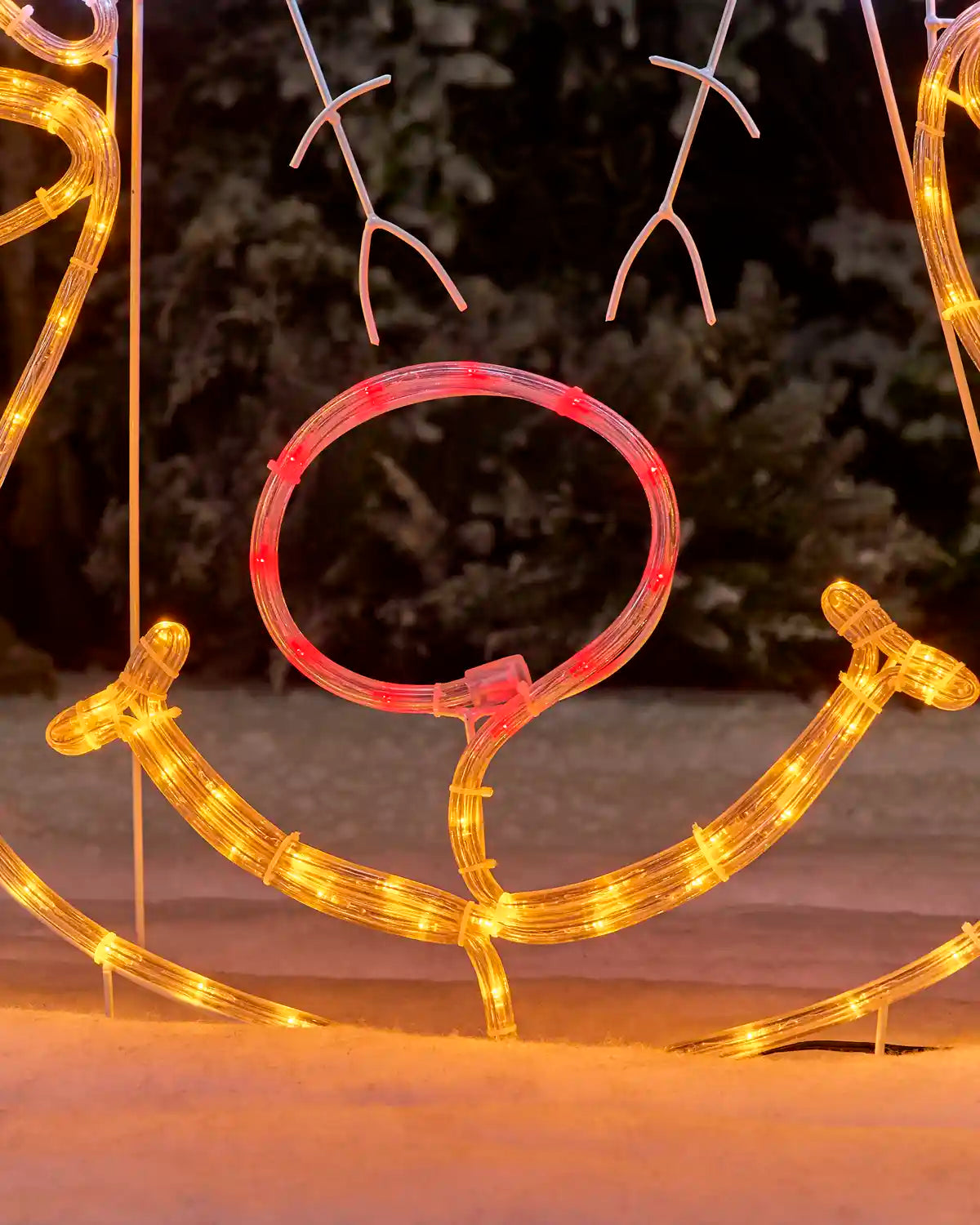 Outdoor holiday light decoration shaped like a reindeer head, with warm yellow wire outlines and a translucent red circular nose ring illuminated with red lights, set in snow at night.