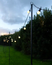 Outdoor light pole holding up festoon lights with a garden background