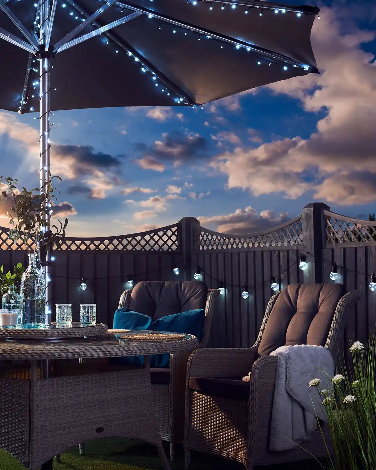 Outdoor patio setting with Icy white string lights wrapped around a large umbrella, table, and chairs under a cloudy sky.
