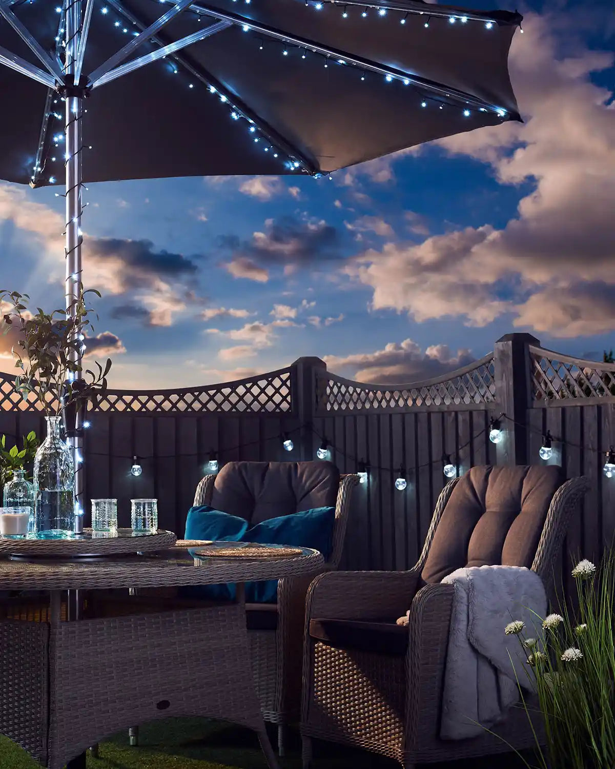 Outdoor patio setting with a large umbrella wrapped in ice white string lights, table, and chairs under a cloudy sky.