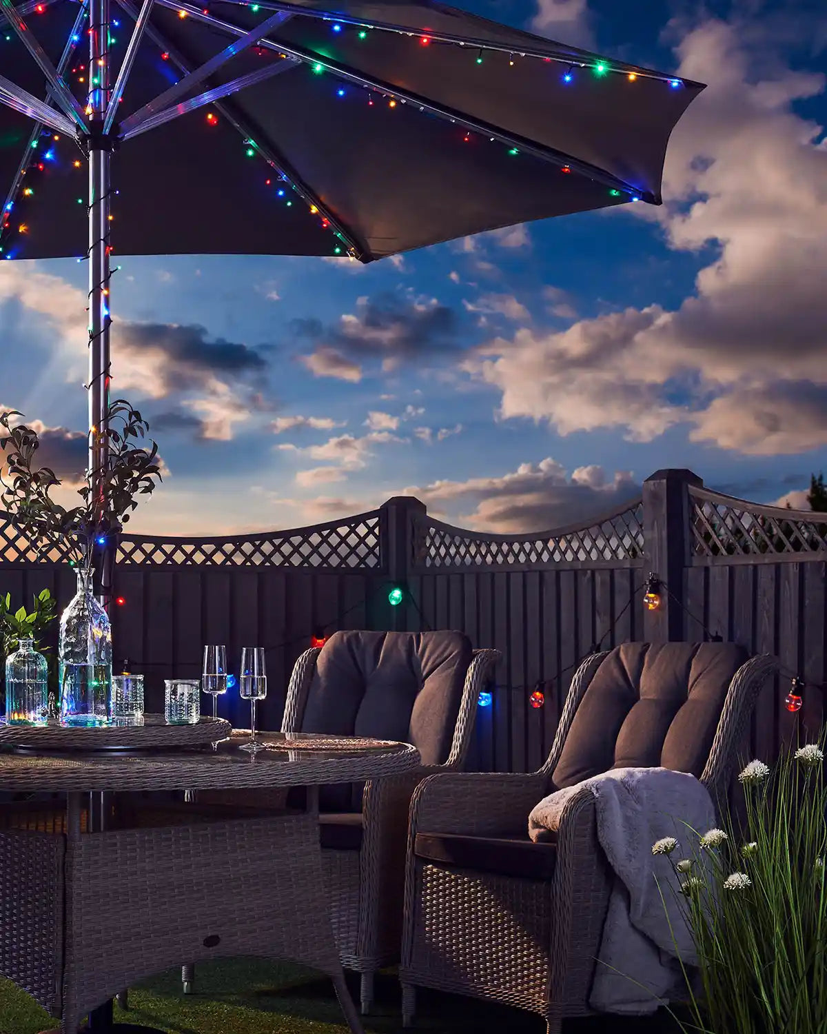 Outdoor patio setting with a lit with multi coloured strong lights on umbrella pole with and table under a cloudy sky
