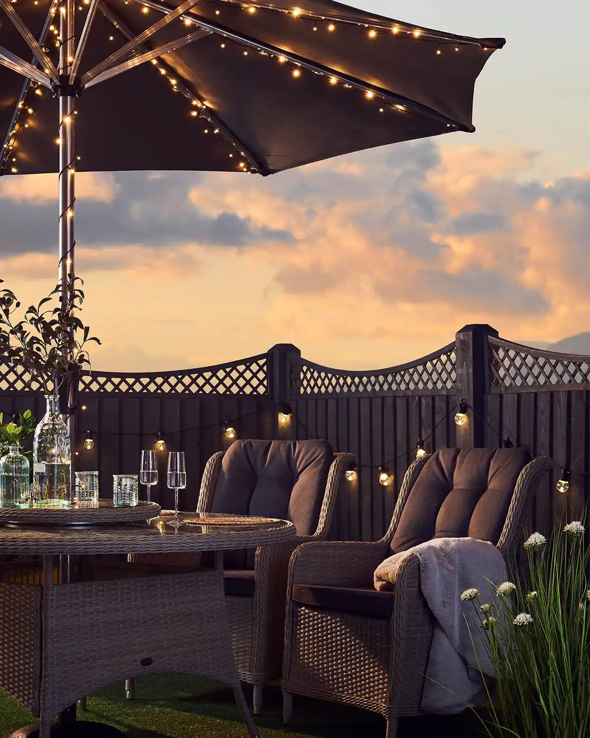 Outdoor patio setting with furniture, warm white string lights wrapped around umbrella pole, and sunset sky