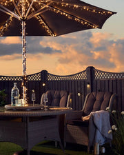 Outdoor patio setting with warm white fairy lights on umbrella, table, and chairs against a sunset sky.