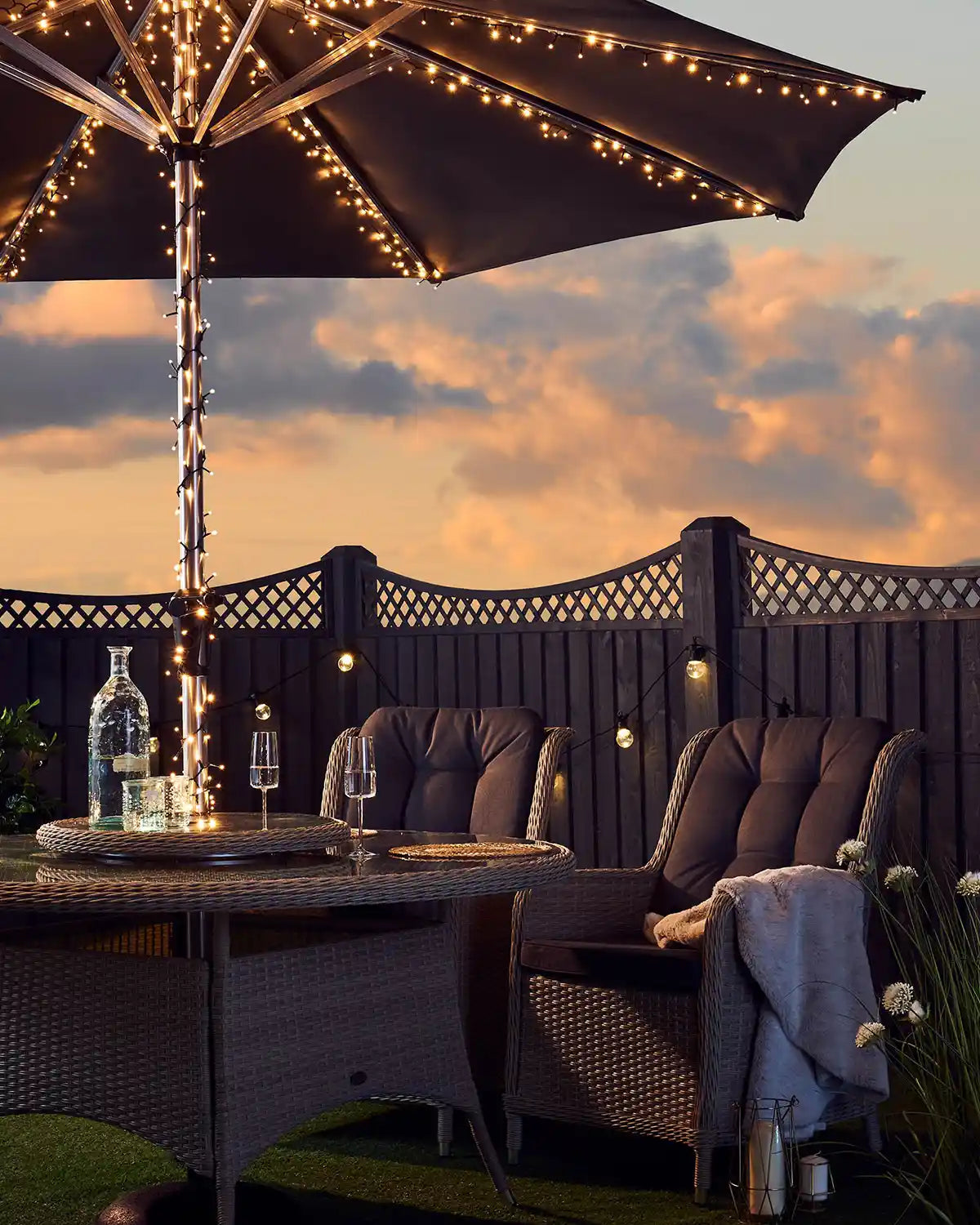 Outdoor patio setting with warm white fairy lights on umbrella, table, and chairs against a sunset sky.