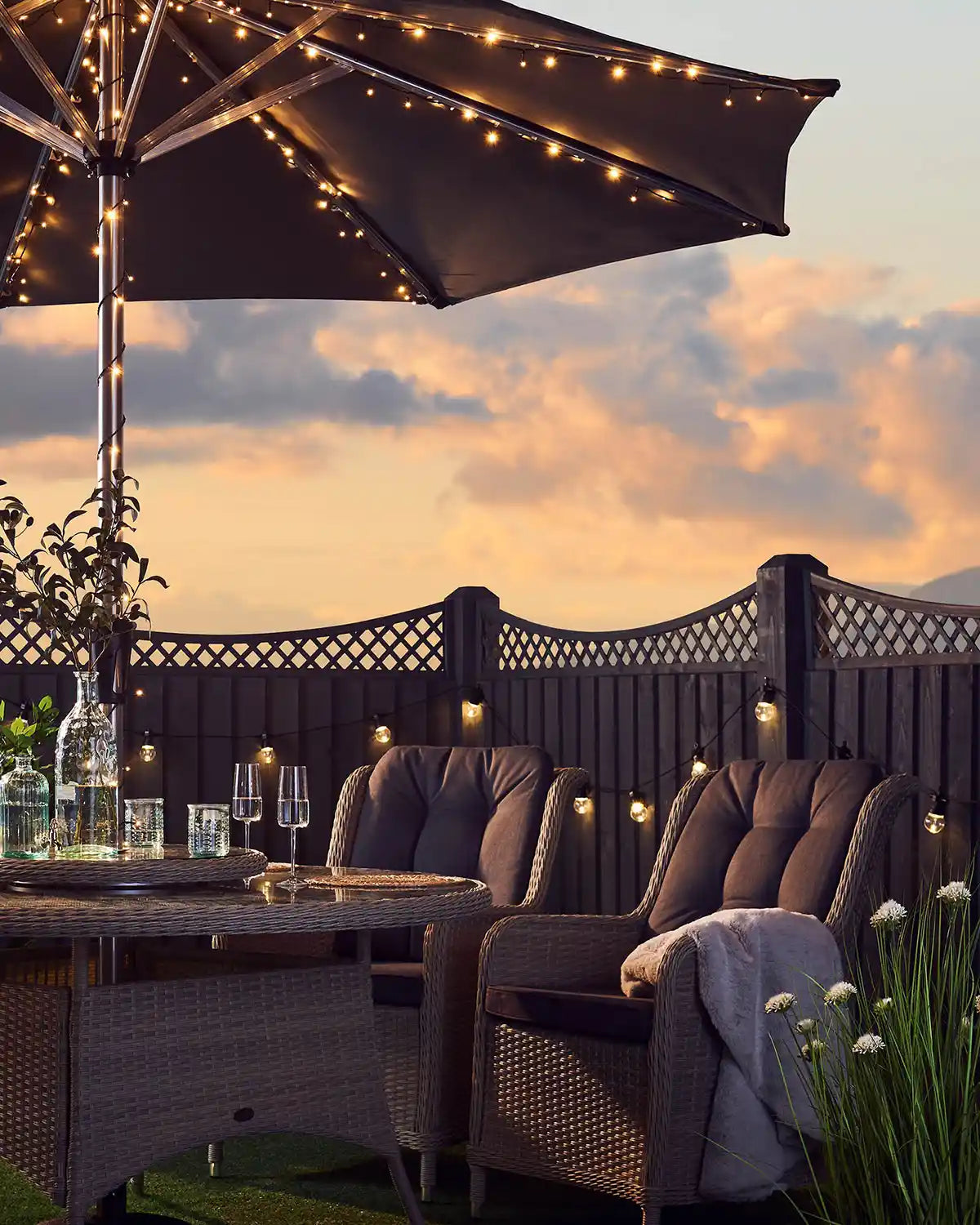 Outdoor patio setting with warm white string lights on an umbrella pole, table, and chairs against a sunset sky.