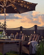 Outdoor patio setting with warm white string lights on an umbrella pole, table, and chairs against a sunset sky.