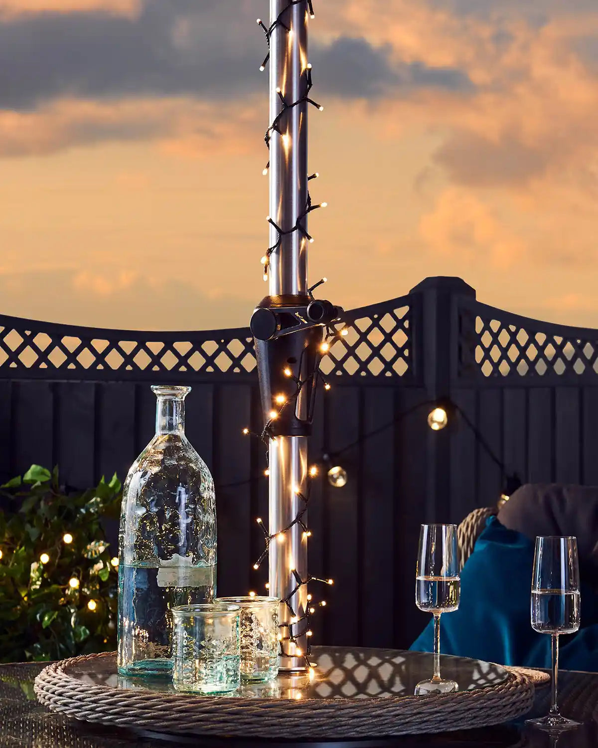 Outdoor setting with a bottle of water, warm white string lights on umbrella pole, glasses, and string lights against a sunset sky.