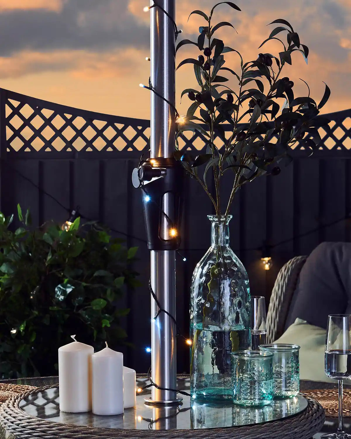 Outdoor setting with a metal pole with warm white and ice white candles, and a vase with plants against a sunset sky.