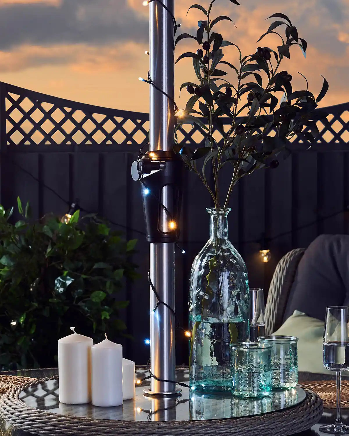 Outdoor setting with a table featuring a vase, warm white and ice white string lights, candles, and glasses against a sunset background