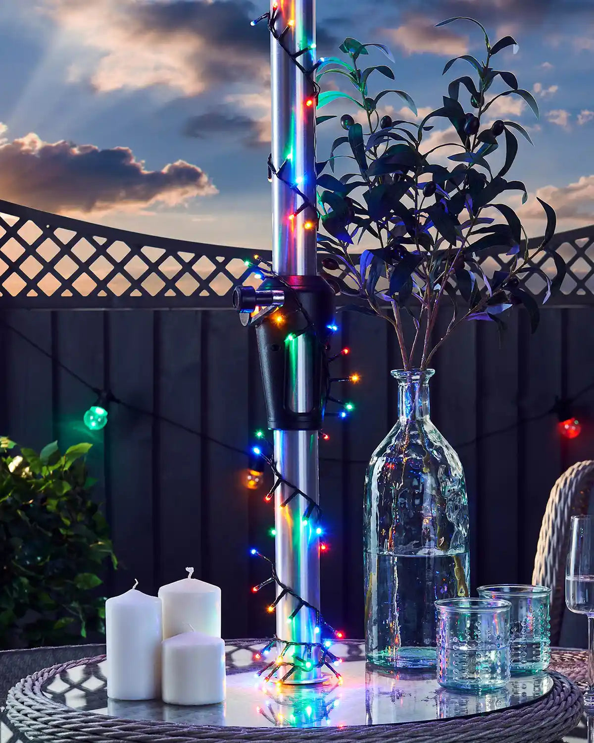 Outdoor setting with a table featuring candles, a vase, and colourful string lights against a sunset sky.