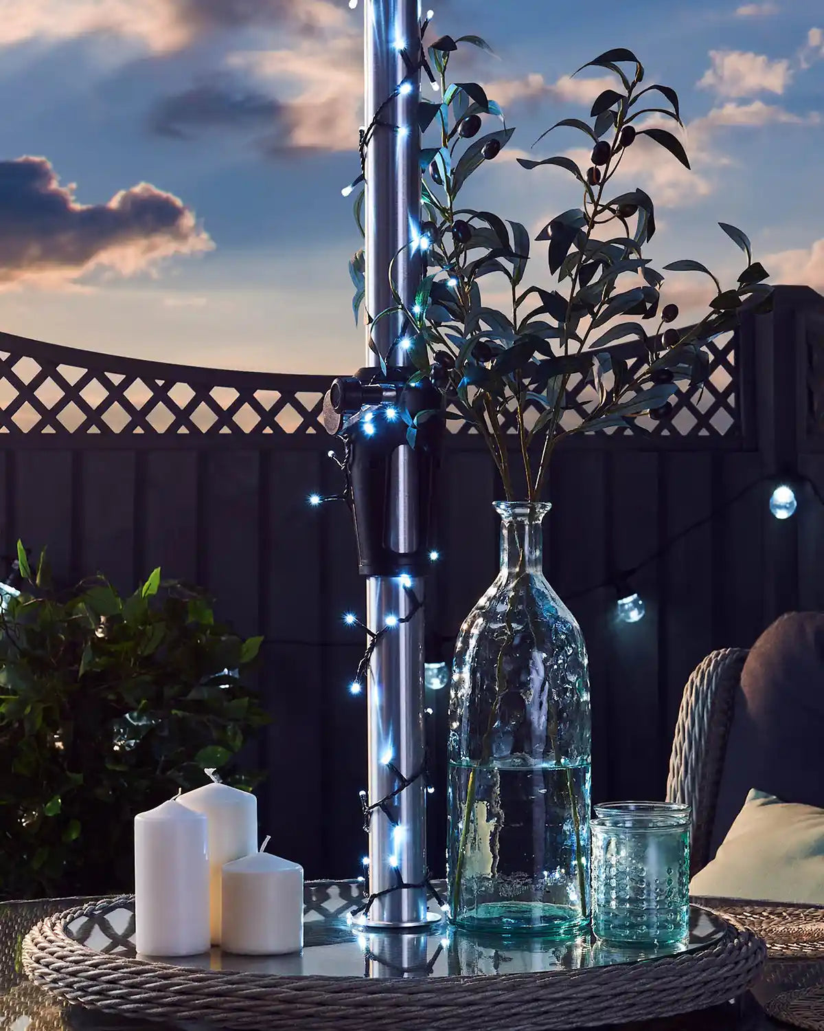 Outdoor setting with a table featuring candles, a vase, and decorative plants against a blue cloudy sky.