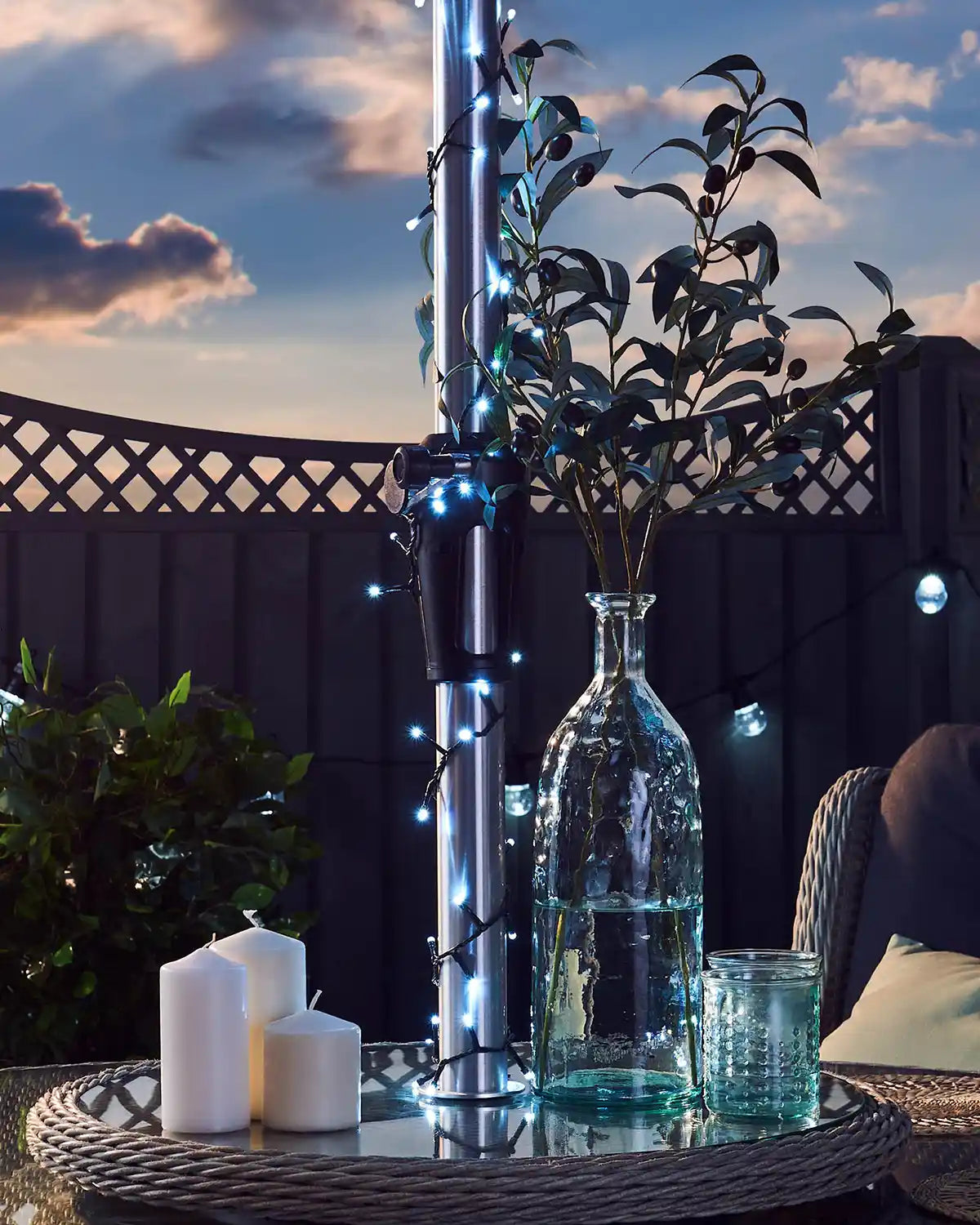 Outdoor setting with a table featuring candles, a vase, and decorative plants and ice white fairy lights against a cloudy blue sky
