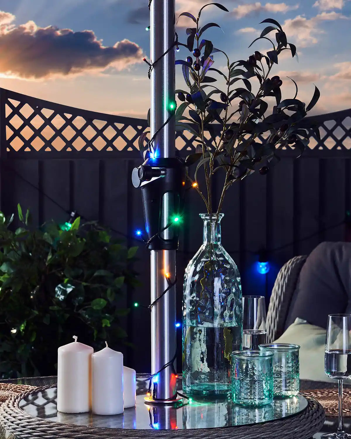Outdoor setting with a table featuring candles, a vase with plants, and colourful string lights against a sunset sky.