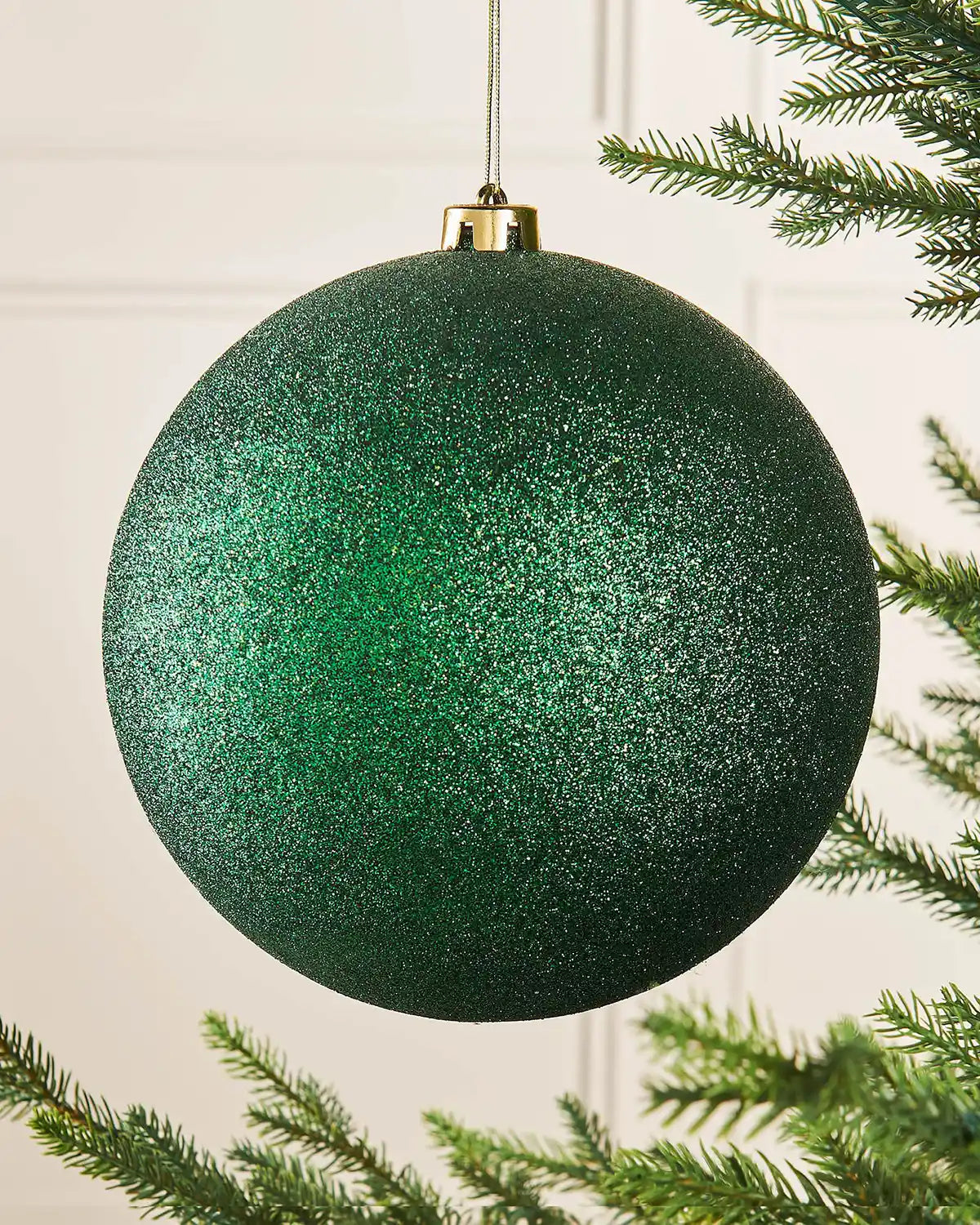 Oversized Christmas Green glittery Christmas ball ornament with a gold hook on a Christmas tree branch against a cream panel background