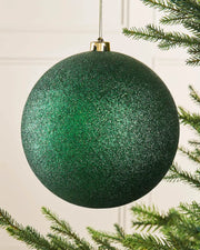 Oversized Christmas Green glittery Christmas ball ornament with a gold hook on a Christmas tree branch against a cream panel background